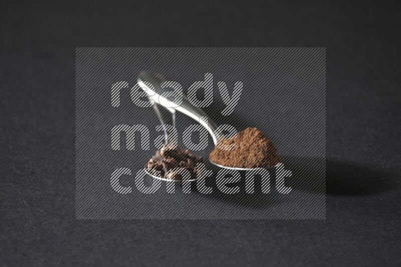 2 metal spoons, one full of cloves grains and the other full of cloves powder on a black flooring