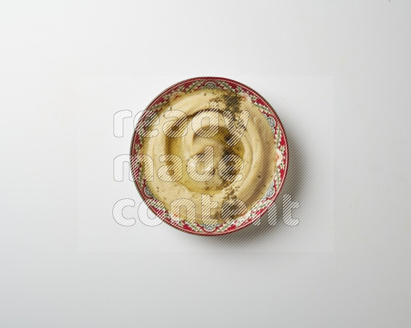 Hummus in a red plate with patterns garnished with zaatar & sumak on a white background