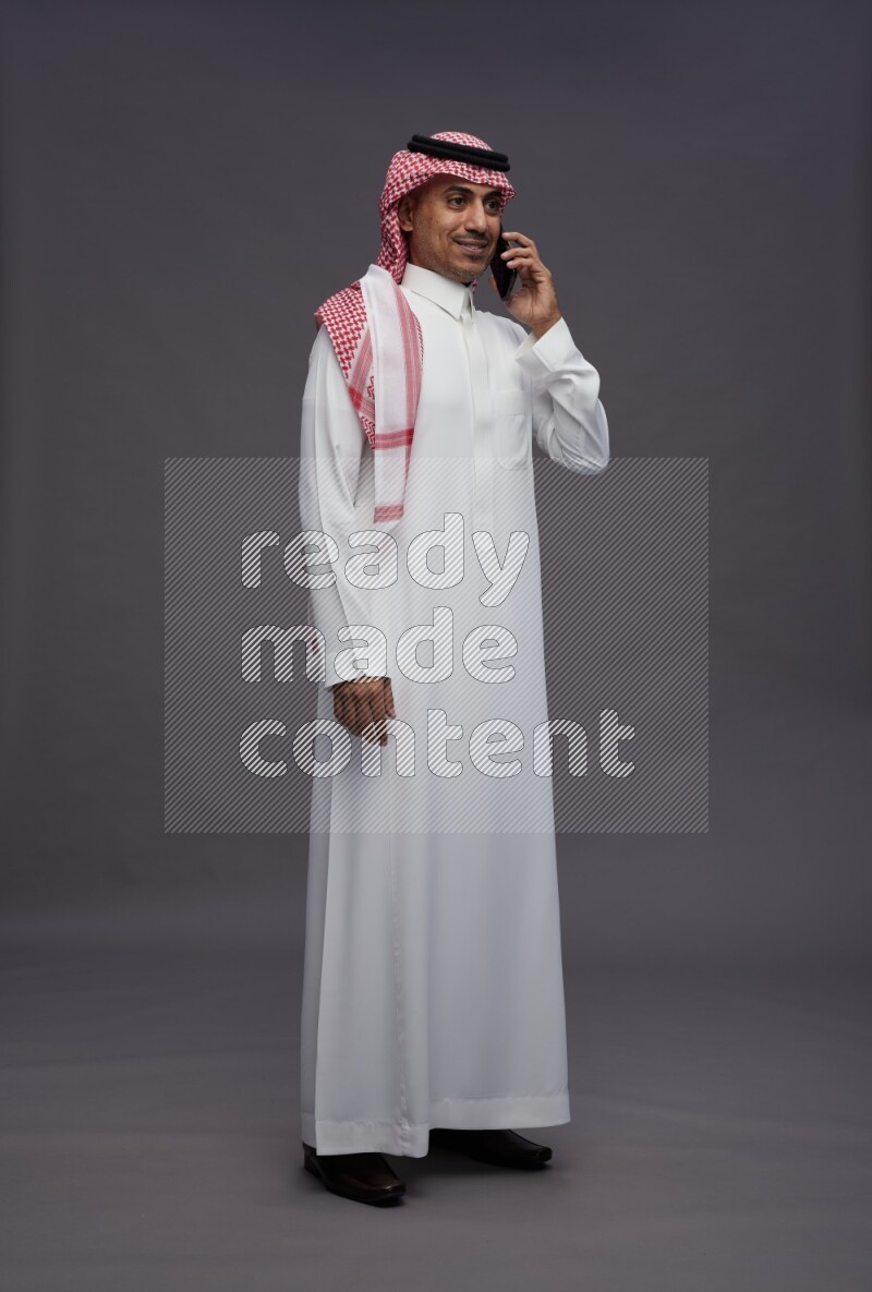 Saudi man wearing thob and shomag standing talking on phone on gray background