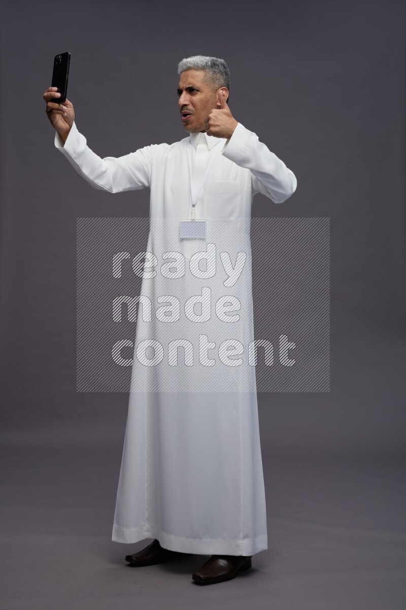 Saudi man wearing thob with neck strap employee badge standing taking selife on gray background
