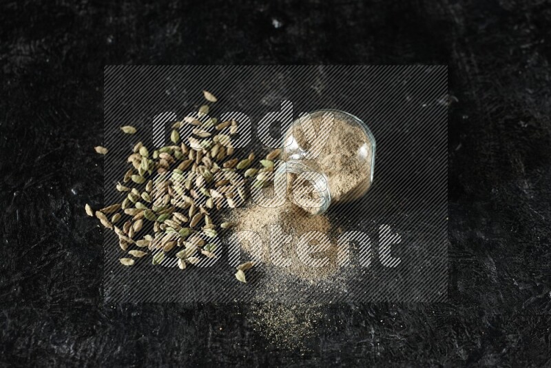 A flipped glass spice jar full of cardamom powder and powder spilled out of it with cardamom seeds spreaded on textured black flooring