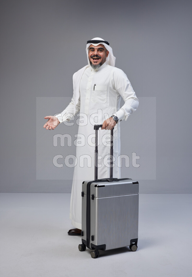 Saudi man wearing Thob and white Shomag standing holding Travel bag on Gray background