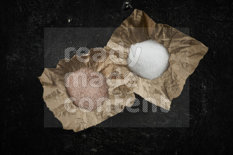 2 crumpled pieces of paper full of pink himalayan salt and white salt on black background