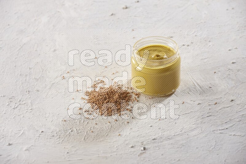 A glass jar full of mustard paste with mustard seeds spread next to it on a textured white flooring