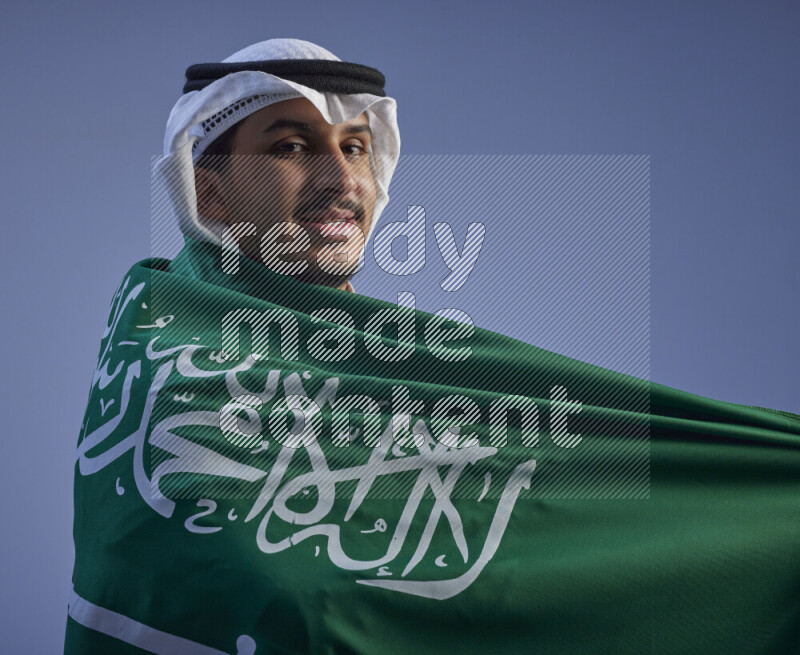 A close-up shot of Saudi man wearing thob and white shomag wrapping big Saudi flag on gray background