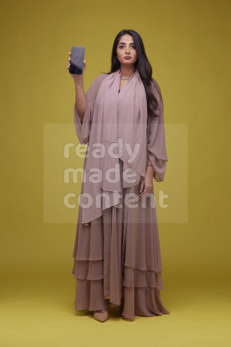 A woman Showing SmartPhone Screen on a Yellow Background wearing Brown Abaya