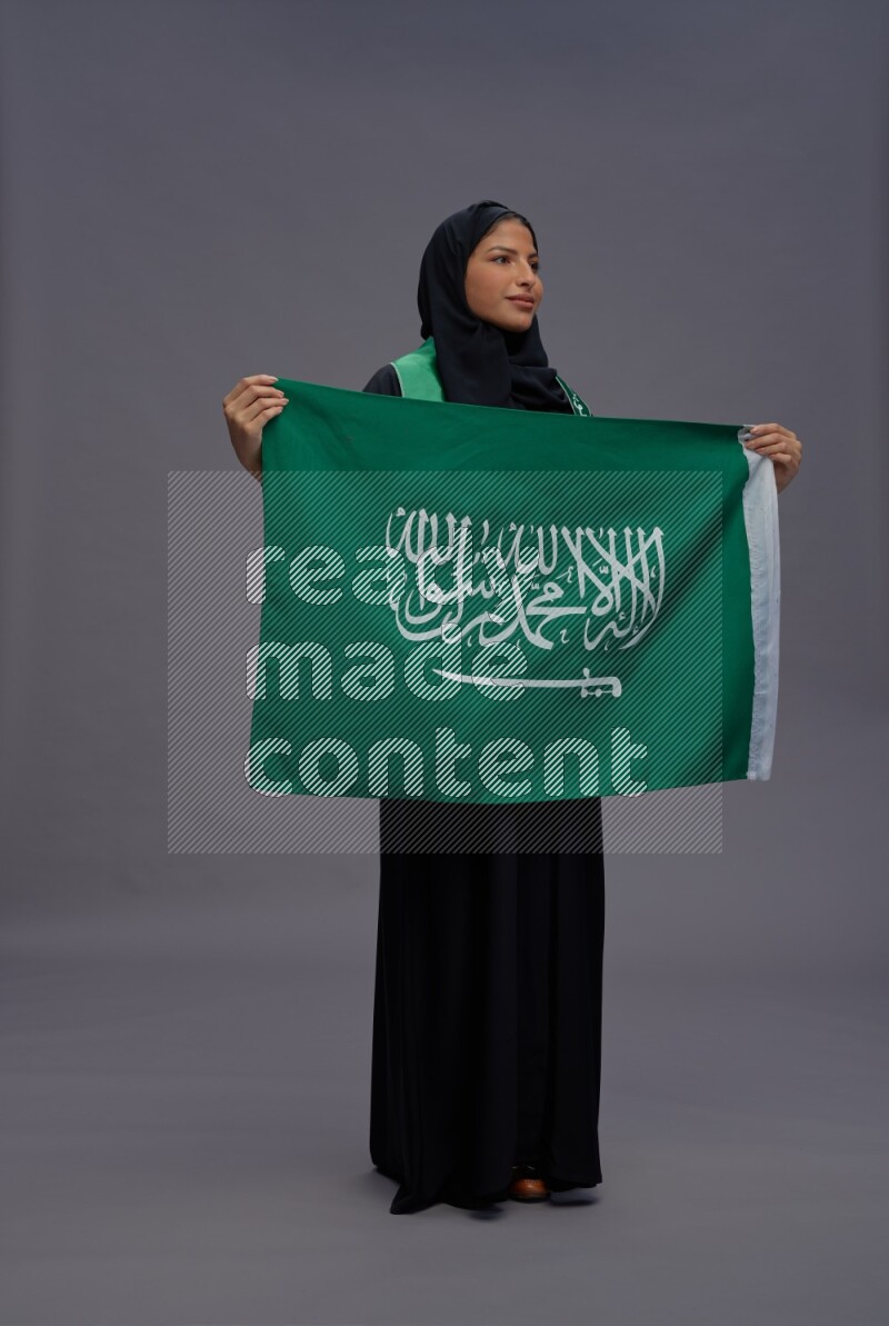 Saudi woman wearing Abaya standing holding Saudi flag on gray background